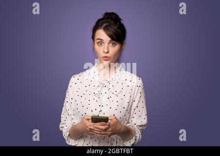 Schockierte Frau hält ein Telefon an einer blauen Studiowand und schaut in einem schönen Kleid auf die Kamera Stockfoto