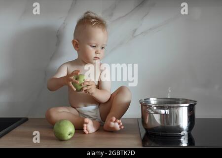 Der kleine Junge sitzt mit grünen Äpfeln auf der Küchenarbeitsfläche und schaut auf den Topf Stockfoto