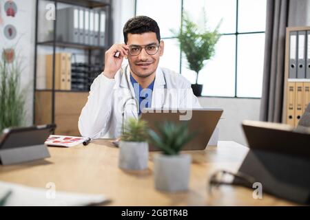 Arabischer Arzt oder Medizinstudent, der im modernen Büro an einem Tisch sitzt und mit einem Laptop arbeitet. Junger, hübscher indischer Arzt, der bei der Arbeit einen Laptop benutzt und seine Brille justiert Stockfoto