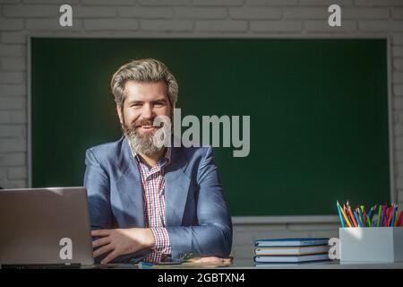 Porträt eines selbstbewussten männlichen Lehrers im Klassenzimmer. Junger männlicher Lehrer mit Laptop in der Klasse sitzt am Tisch an der Tafel im Klassenzimmer. Lehrer Stockfoto