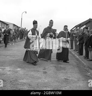 In den 1960er Jahren versammeln sich historische Einheimische in Lochgelly, einer kleinen Bergbaustadt in Fife, Schottland, draußen auf dem Bürgersteig, um den Priestern in ihren Kittel Respekt zu erweisen, die auf ihrem Weg zur Weihe in der Kirche der Gemeinde vorbei gehen. Stockfoto