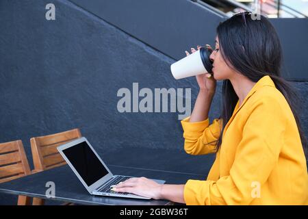 Junge lateinische Geschäftsfrau, die mit einem Laptop arbeitet und Kaffee im Freien trinkt. Stockfoto
