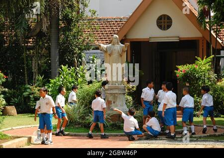 Eine Gruppe von Grundschülerinnen und Grundschülerinnen spielt zusammen um eine Jesus-Statue vor einer Kapelle. Stockfoto