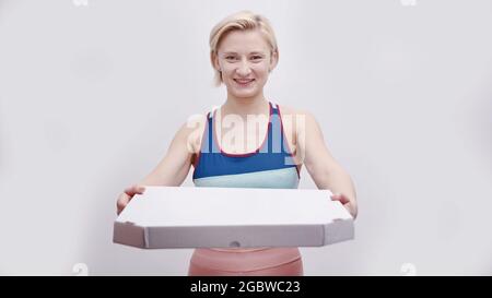 Mädchen in aktiver Kleidung mit Pizza-Box. Verschieben der Kamera. Konzept der Wahl zwischen einem gesunden Lebensstil und Junk-Food. Sportliche Mädchen hält Pizza isoliert auf weißem Hintergrund. Stockfoto