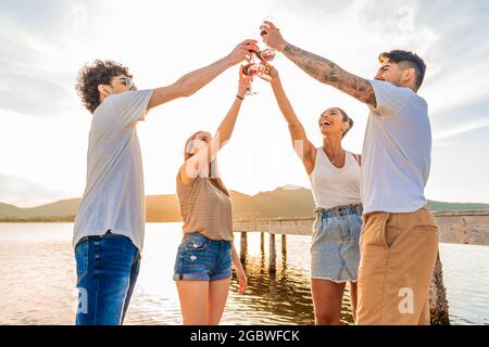 Gruppe von multirassischen glücklichen Freunden, die bei Sonnenuntergang am Strand mit Rotwein toasten und vor Sonnenlicht in hinterleuchteter Beleuchtung Gläser klirren. Junge Menschen haben Stockfoto