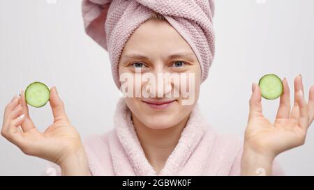 Mädchen mit einem Kopftuch, das einen rosa Bademantel trägt und Gurkenscheiben in den Händen hält. Konzept der Verschönerung und Hautpflege während des Aufenthalts zu Hause. Mädchen in guter Laune verwöhnen sich. Stockfoto