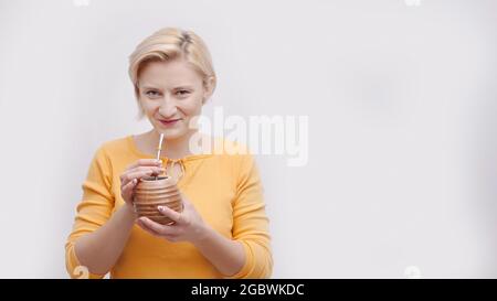 Schönes Mädchen, das eine braune Behälterform wie eine Kokosnuss mit einem Stroh hält. Im Begriff, mit dem Stroh zu trinken. Das Mädchen ist in einem gelben Oberteil gekleidet. Isoliert auf weißem Hintergrund. Tropischer Urlaub. Stockfoto
