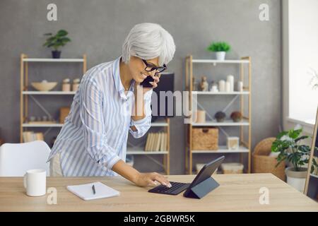 Glückliche, reife Geschäftsfrau, die im Heimbüro arbeitet, mit dem Mobiltelefon spricht und ein Tablet benutzt Stockfoto