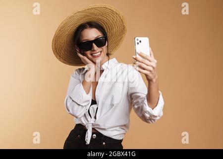 Junge Frau im Strohhut lächelt, während sie Selfie auf dem Handy gemacht hat isolierter beiger Hintergrund Stockfoto