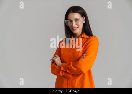 Junge Brünette Frau in Schutzbrille lächelnd und Blick auf die Kamera isoliert auf grauem Hintergrund Stockfoto