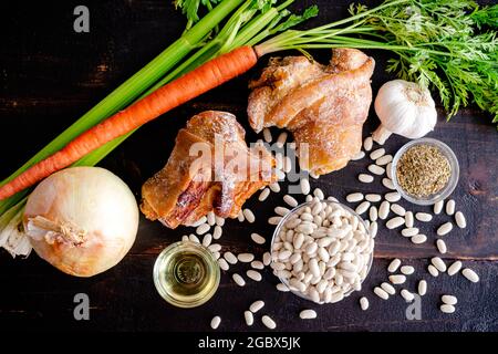 Weiße Bohnen- und Schinkensuppe Zutaten auf dunklem Holz Hintergrund: Geräucherte Schinkenhahne, frisches Gemüse und Gewürze auf einem dunklen Holztisch Stockfoto