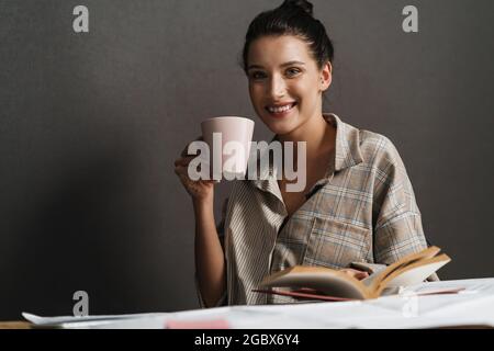 Lächelnd mittleren Alters weiße beiläufige Brünette Frau liest Tagebuch, während sie am Schreibtisch sitzt und Becher hält Stockfoto
