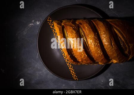 Ungewöhnliches zweifarbiger frischer Laib Brot auf einer Keramikplatte auf schwarzem Hintergrund, Foto in einem niedrigen Ton bei hartem Licht, Nahaufnahme. Stockfoto