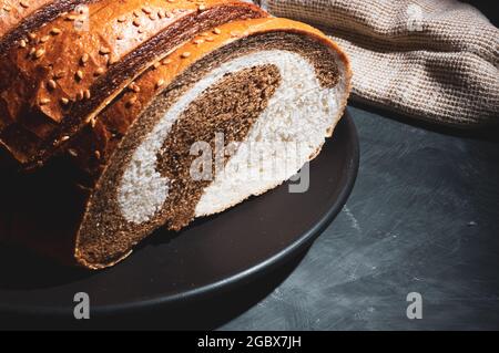 Ungewöhnliches, zweifarbiges frisches Brot in einer Keramikplatte und eine gewebte Serviette auf schwarzem Hintergrund, Foto in einem niedrigen Ton bei hartem Licht, Nahaufnahme. Stockfoto