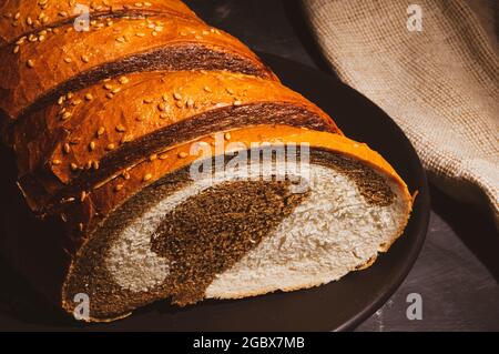 Ungewöhnliches, zweifarbiges frisches Brot in einer Keramikplatte und eine gewebte Serviette auf schwarzem Hintergrund, Foto in einem niedrigen Ton bei hartem Licht, Nahaufnahme. Stockfoto
