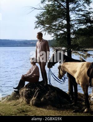 MANN AUS DEN 1930ER JAHREN, DER AUF EINEM FELSEN AM SEE STEHT UND ZWEI PFERDE IN DER HAND HÄLT, WÄHREND EINE FRAU NEBEN IHM SITZT UND VON DER SEITE SCHAUT - H2855C HAR001 HARS FARBE AKTION ALTE ZEIT ZUKUNFT NOSTALGIE ALTE MODE 1 STIL JUNGE ERWACHSENE PFERDE FRIEDEN REITEN REICH LIFESTYLE FRAUEN VERHEIRATET LÄNDLICHEN EHEGATTEN EHEMÄNNER ERWACHSEN GESUNDHEIT LUXUS NATUR SITZEND KOPIE RAUM FREUNDSCHAFT DAMEN PERSONEN INSPIRATION MÄNNER GELASSENHEIT TRANSPORT MÄNNER UND FRAUEN PARTNER SEEUFER AKTIVITÄT TRÄUME ZÜGEL SÄUGETIERE BEOBACHTER ABENTEUER ENTDECKUNG FREIZEIT BEOBACHTER STILE AUFREGUNG ERHOLUNG MANN UND FRAU TRAIL GEHOBENER WOHLSTAND NEBEN ENGLISCHEM SATTEL Stockfoto