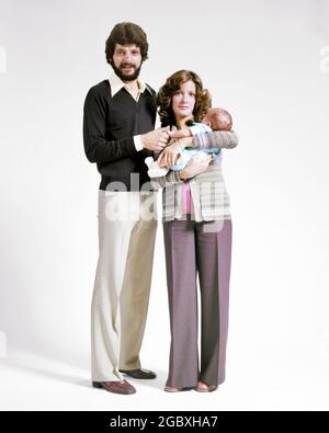 FAMILIENPORTRAIT DER 1970ER JAHRE MANN UND FRAU VATER UND MUTTER STEHEN ZUSAMMEN UND BLICKEN AUF DIE KAMERA MIT EINEM KLEINKIND IN ARMEN - KB12402 HAR001 HARS PAAR NEUGEBORENE FARBE MÜTTER ALTE ZEIT NOSTALGIE ALTE MODE BART 1 JUGENDLICH STIL JUNGEN ERWACHSENEN KLEINKIND STARKE SÖHNE FAMILIEN FRAUEN VERHEIRATET EHEPARTNER EHEMÄNNER DAMEN PERSONEN FÜRSORGLICH MÄNNER VÄTER PARTNER SCHNURRBART SCHNURRBART UND VÄTER IM GESICHTSHAAR BABY JUNGE PERSÖNLICHE BINDUNG ZUNEIGUNG BÄRTE EMOTION JUGENDLICHE MÜTTER FRAUEN JUNGER ERWACHSENER MANN JUNGER ERWACHSENER FRAU KAUKASISCHE ETHNIE HAR001 ALT MODISCH Stockfoto