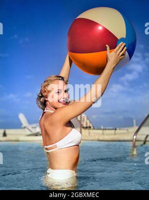 1950 SEITENANSICHT DER BLONDINE IN WEISSEM BIKINI, DER IM SCHWIMMBAD IM HÜFTHOHEN WASSER STEHT UND DEN STRANDBALL IN DER LUFT HÄLT - S886C HAR001 HARS GESUNDHEIT KOPIEREN RAUM HALBE LÄNGE BLICKKONTAKT ZEIT WEG GLÜCK SCHWIMMBAD FREIZEIT REISE KURZURLAUB ERHOLUNG ERHOLUNG SCHWIMMEN URLAUB LÄCHELT SCHWIMMER ZWEITEILIGE STILVOLLE BADEANZUG SCHWIMMEN TRAGEN FERIEN TAILLE-HOCH JUNGE ERWACHSENE FRAU KAUKASISCHE ETHNIE HAR001 ALTMODISCH Stockfoto