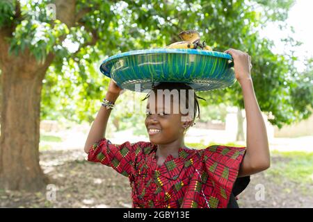 Auf diesem Bild flirtet ein wunderschön gekleidetes, anmutiges kleines afrikanisches Mädchen, das einen Teller Bananen auf dem Kopf trägt, mit der Kamera Stockfoto