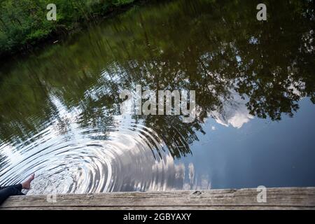 Die Füße baumeln im Waldteich Wasser von einem hölzernen Dock, was kreisförmige Wellen macht Stockfoto