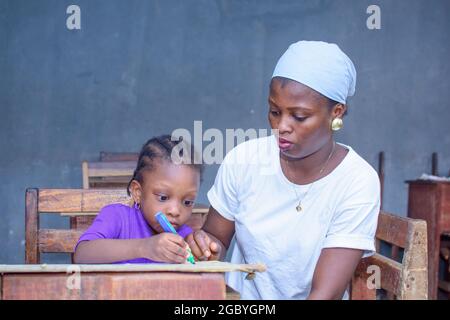 Afrikanische nigrische Mutter oder Lehrerin, die mit ihrem Kind in einem Klassenzimmer sitzt und ihr beim Studium hilft, in ihrer Schule hervorragende Leistungen zu erzielen Stockfoto