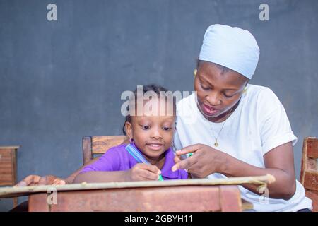 Afrikanische nigrische Mutter oder Lehrerin, die mit ihrem Kind in einem Klassenzimmer sitzt und ihr beim Studium hilft, in ihrer Schule hervorragende Leistungen zu erzielen Stockfoto