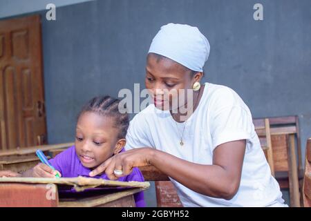 Afrikanische nigrische Mutter oder Lehrerin, die mit ihrem Kind in einem Klassenzimmer sitzt und ihr beim Studium hilft, in ihrer Schule hervorragende Leistungen zu erzielen Stockfoto