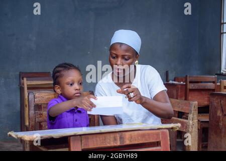 Afrikanische nigrische Mutter oder Lehrerin, die mit ihrem Kind in einem Klassenzimmer sitzt und ihr beim Studium hilft, in ihrer Schule hervorragende Leistungen zu erzielen Stockfoto