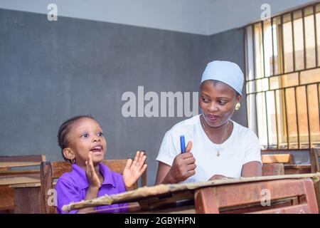 Afrikanische nigrische Mutter oder Lehrerin, die mit ihrem Kind in einem Klassenzimmer sitzt und ihr beim Studium hilft, in ihrer Schule hervorragende Leistungen zu erzielen Stockfoto