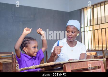 Afrikanische nigrische Mutter oder Lehrerin, die mit ihrem Kind in einem Klassenzimmer sitzt und ihr beim Studium hilft, in ihrer Schule hervorragende Leistungen zu erzielen Stockfoto