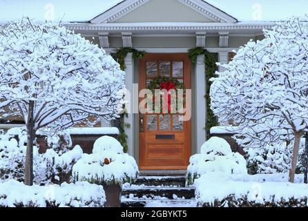 Hölzerne Eingangstür mit Weihnachtskranz, umgeben von schneebedeckten Büschen und Bäumen Stockfoto