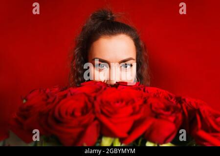 Porträt eines Mädchens, das hinter Blumenstrauß auf rotem Hintergrund herausguckt. Portrait des Mädchens späht aus hinter Blumenstrauß auf rotem Hintergrund. Junge Frau mit großem Strauß roten Rosen Stockfoto