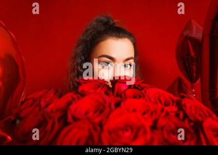 Porträt eines Mädchens, das hinter Blumenstrauß auf rotem Hintergrund herausguckt. Portrait des Mädchens späht aus hinter Blumenstrauß auf rotem Hintergrund. Junge Frau mit großem Strauß roten Rosen Stockfoto