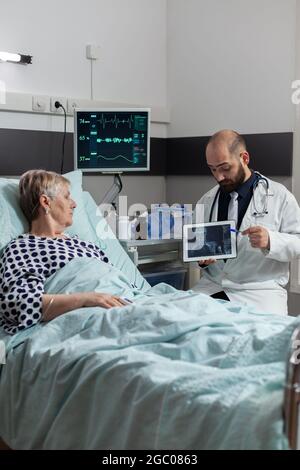 Kranke ältere Frau, die ein Medikament über eine intravenöse Leitung erhält, liegt im Krankenhauszimmer und hört dem Arzt zu, wie er über Körpertrauma spricht, das auf digitale Röntgenaufnahmen auf dem Tablet-pc zeigt. Stockfoto