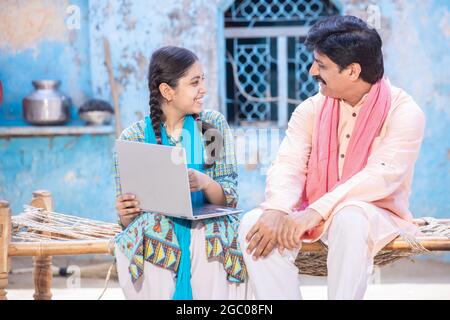 Fröhlicher indischer Vater und junge Tochter, die einen Laptop benutzen und sich gegenseitig anblicken, während sie vor ihrem Haus auf einem traditionellen Holzbett sitzen, Happy Rural Stockfoto