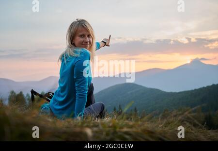 Mädchen mit einem leichten Lächeln im Gesicht, das auf einem Hügel ruht, das Panorama der Berggipfel genießt und ihre Hand auf die Wolken am Abendhimmel bei der Sonne zeigt Stockfoto