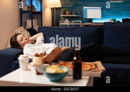 Kaukasische Frau schläft ein, nachdem sie Junk-Food im Wohnzimmer spät in der Nacht gegessen hat, während sie Essen zum Mitnehmen nach Hause geliefert hat. Leckere köstliche Mahlzeit Mittagessen Bestellung. Ungesunde Snacks Fastfood Stockfoto