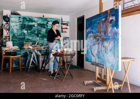 Konzentrierte Künstlerin mittleren Alters, die blaue Farbe mischte und Geräte in ihrem Home Art Studio verwendete. Stockfoto