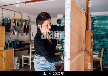 Fokussierte Malerin, die sich in ihrer Garage mit blauer Farbe auf einer großen Leinwand entspannt. Stockfoto