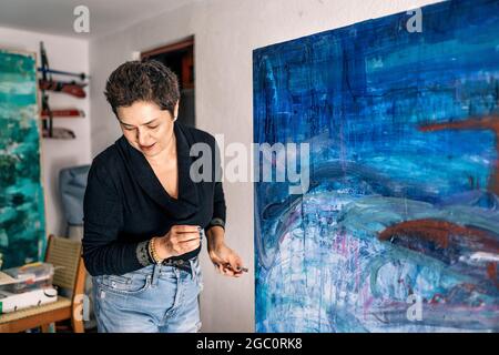 Glückliche Malerin, die sich in ihrer Garage mit blauer Farbe auf einer großen Leinwand entspannt. Stockfoto