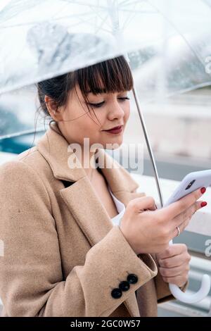 Eine lächelnde junge Frau, die an regnerischen Tagen mit ihrem Mobiltelefon einen Regenschirm auf der Straße hält. Stockfoto