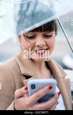 Eine lächelnde junge Frau, die an regnerischen Tagen mit ihrem Mobiltelefon einen Regenschirm auf der Straße hält. Stockfoto