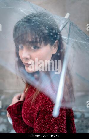Schöne Frau mit transparentem Regenschirm und Blick auf die Kamera. Sie trägt legere Kleidung. Stockfoto