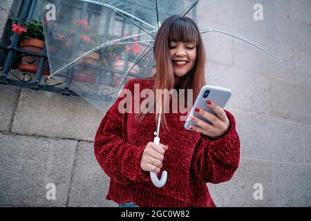 Junge Frau, die an regnerischen Tagen einen transparenten Regenschirm verwendet, ihr Mobiltelefon auf der Straße benutzt. Stockfoto