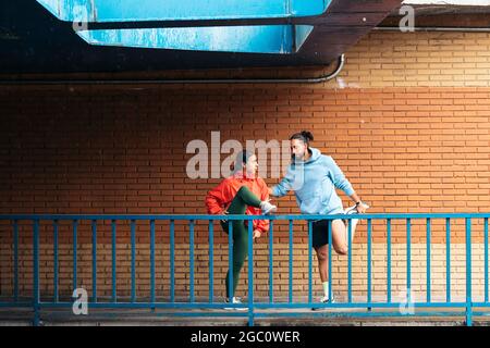 Glückliche Frau und ihre Freundin tragen Sportkleidung, die sich nach dem Laufen in der Stadt an regnerischen Tagen dehnt. Sie lächeln und reden. Stockfoto