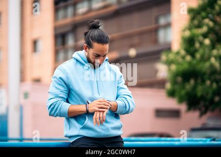 Schöner Mann mit Sportkleidung, der seine Smart-Watch verwendet und sich nach dem Laufen in der Stadt ausruhte. Stockfoto