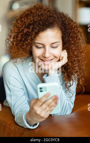Junge, lockig rothaarige Frau, die am Telefon chattet und weit lächelt, sich zu Hause auf einem bequemen Sofa niederlegt, Nachrichten von der Familie erhielt, Zeit in sozialen Netzwerken verbringt, vertican shot Stockfoto