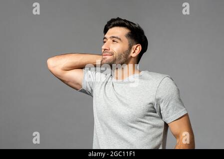 Porträt eines jungen Mannes, der mit der Hand hinter dem Hals lächelt, wegschaut und nach dem Aufwachen nachdenkt, aufgenommen im Studio auf einem isolierten hellgrauen Hintergrund Stockfoto