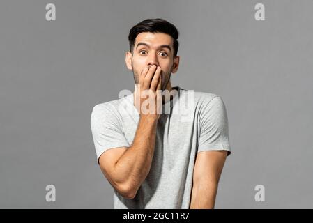 Porträt eines schockierten jungen kaukasischen Mannes mit einem handbedeckenden Mund auf hellgrauem Studiohintergrund Stockfoto