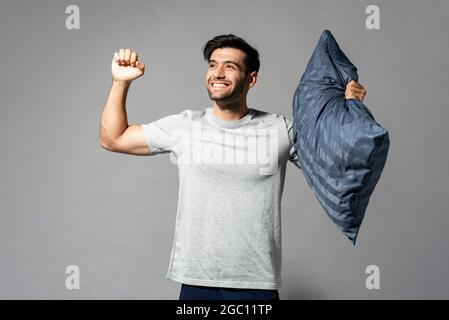 Lächelnder, frischer, lebhafter junger Mann, der Kissen hält und sich nach einer guten Aufnahme im Schlafstudio auf einem isolierten hellgrauen Hintergrund dehnt Stockfoto